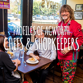 A smiling man in a red shirt stands next to a seated couple in a restaurant. The text overlay reads, "Profiles of Acworth - Chefs & Shopkeepers.