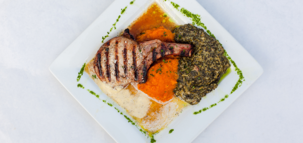 A grilled pork chop served with a side of mashed sweet potatoes and spinach, plated with sauce and garnished with green herbs on a white square plate.