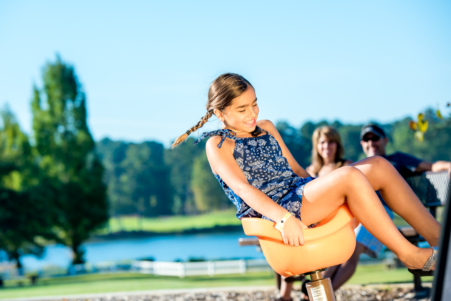A girl with braided hair spins on a playground apparatus outdoors, smiling, as two adults relax nearby by the lake—a joyful moment at one of the Best Playgrounds in Atlanta.