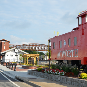 A red train caboose labeled "Acworth" sits near a small white building, a clock, flowers, and a pedestrian bridge with "Acworth" written on it—capturing the charm of Acworth travel.