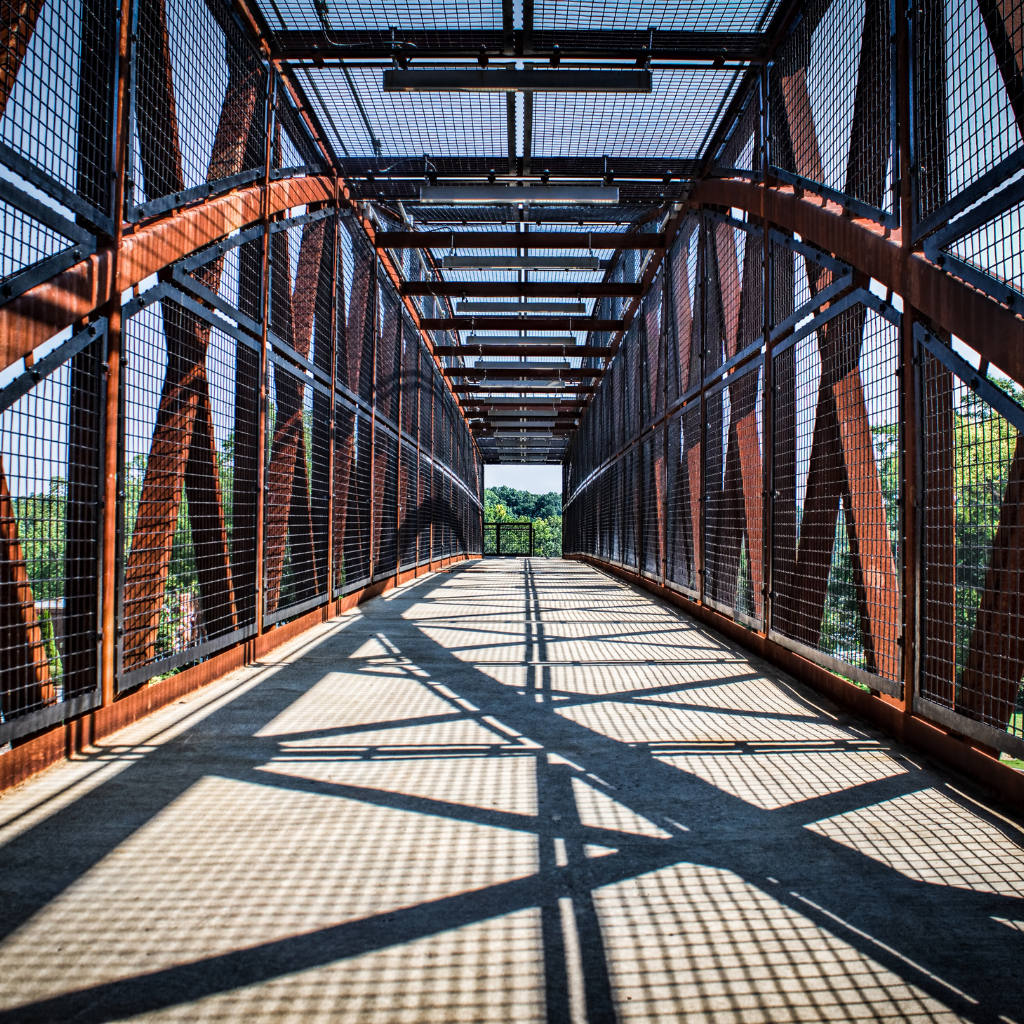Pedestrian bridge with metal fencing and red beams casts a grid-like shadow on the concrete walkway, with trees visible in the background—a unique sight during Acworth travel.