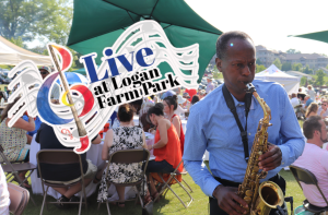 A man plays saxophone at an outdoor event with seated attendees under tents, with a "Live at Logan Farm Park" graphic in the foreground.