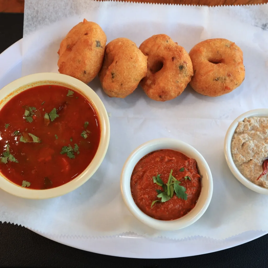 A plate with four medu vada, served with bowls of sambar, red chutney, and white coconut chutney on a sheet of parchment paper.