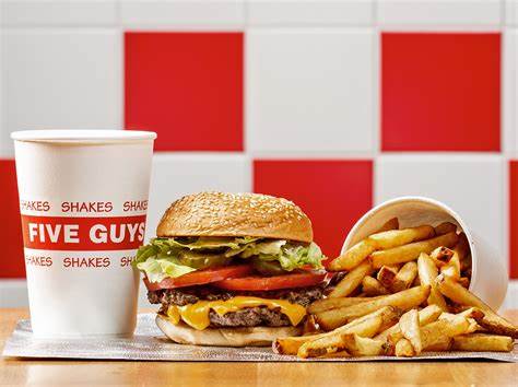 A Five Guys meal with a cheeseburger topped with lettuce and tomato, a cup of fries, and a drink in front of a red and white checkered wall.