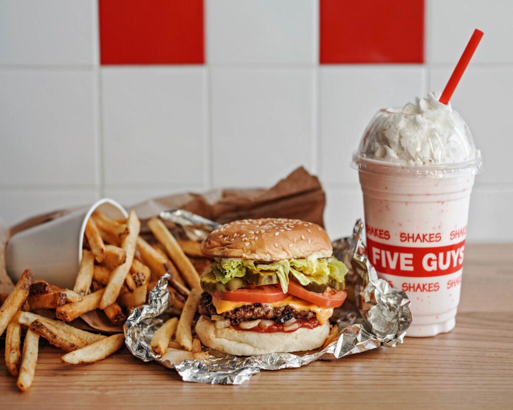 Burger with lettuce and tomato on foil, fries in a cup, and a whipped cream-topped shake in a branded cup with a straw on a wooden table.