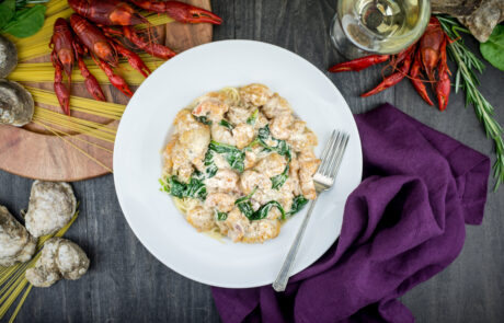 A plate of creamy pasta with shrimp and spinach, surrounded by crayfish, oysters, uncooked spaghetti, and a glass of white wine. A purple cloth is beside the plate.