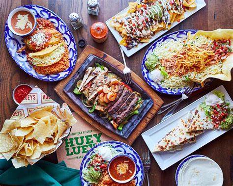 A table filled with Mexican dishes including nachos, fajitas, chips with salsa, and more. Plates are on a wooden surface with condiments and menu visible.