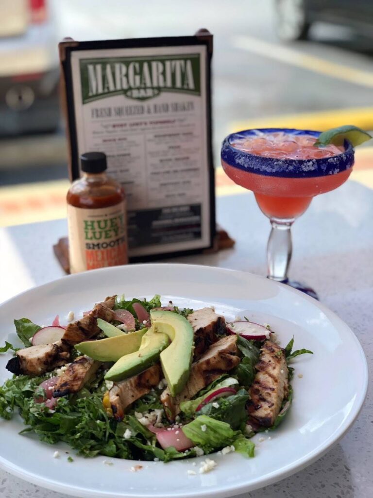 A plate of grilled chicken salad with avocado slices and a strawberry margarita in a glass with a salted rim. Hot sauce and a menu are in the background.