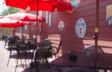 Outdoor patio with metal tables and chairs, under red umbrellas. Wall nearby decorated with various signs. Text reads "Perfect Patio Weather.