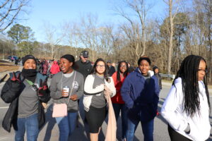 A group of people walking together on a paved path on a sunny day; trees and sparse vegetation are in the background.