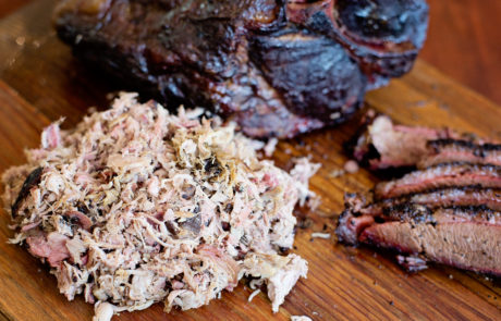 Shredded and sliced smoked meat on a wooden cutting board, with a large piece of smoked meat in the background.