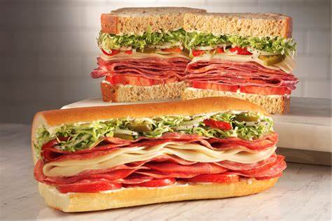 Two deli sandwiches with multiple layers of meats, cheese, shredded lettuce, and tomatoes on different types of bread.