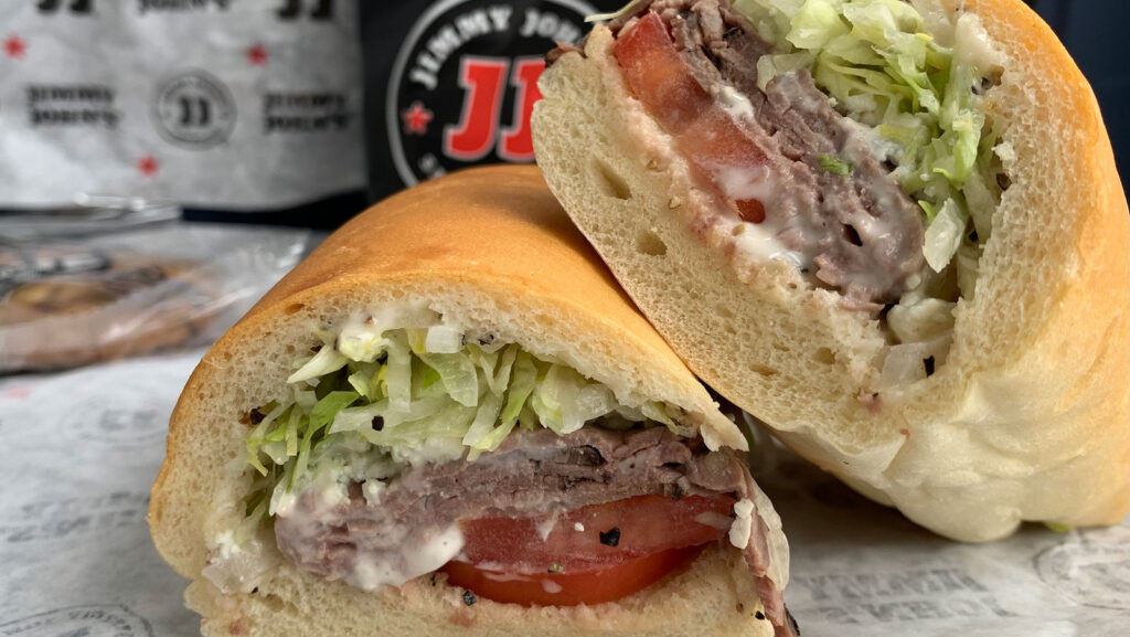 A sub sandwich with sliced roast beef, lettuce, tomato, and mayonnaise is cut in half. It's placed on a wrapper, with branded packaging in the background.