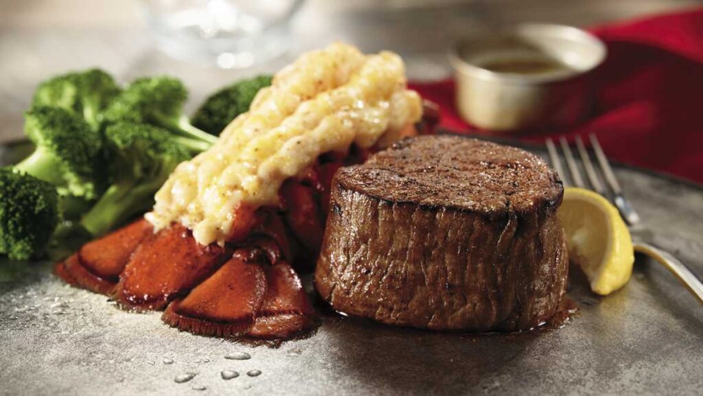 A grilled steak and lobster tail served with broccoli, a lemon wedge, and a sauce dish on a plate.