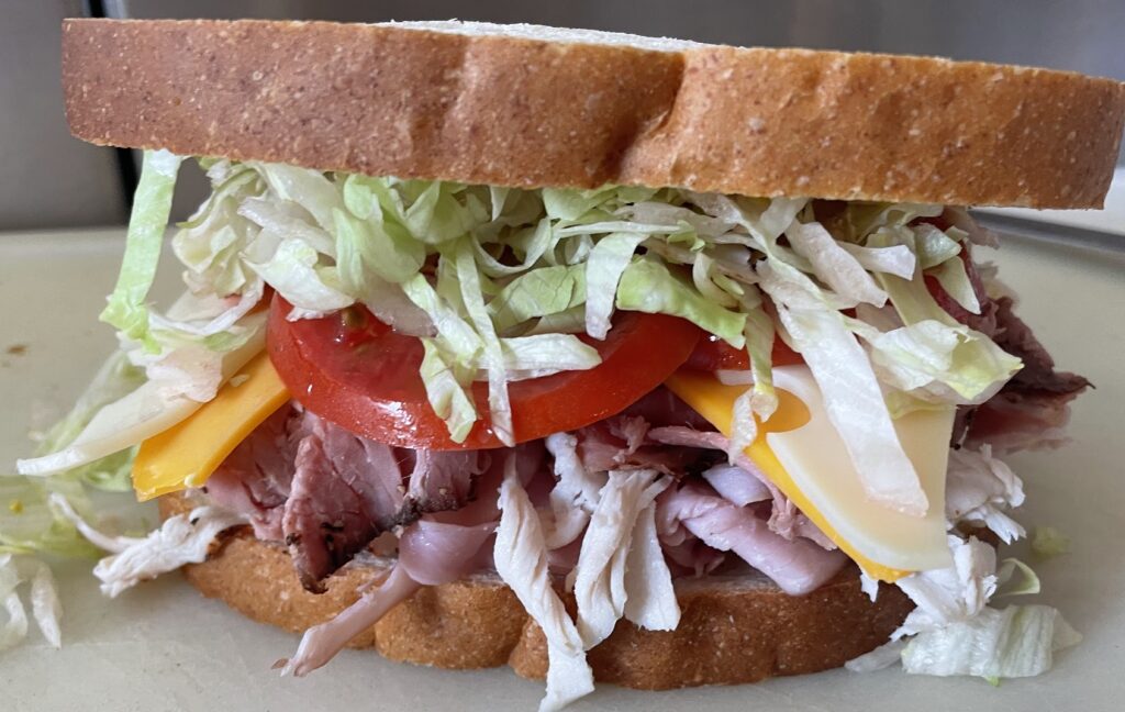 A sandwich with roast beef, cheddar, tomato slices, shredded lettuce, and white bread, placed on a cutting board.