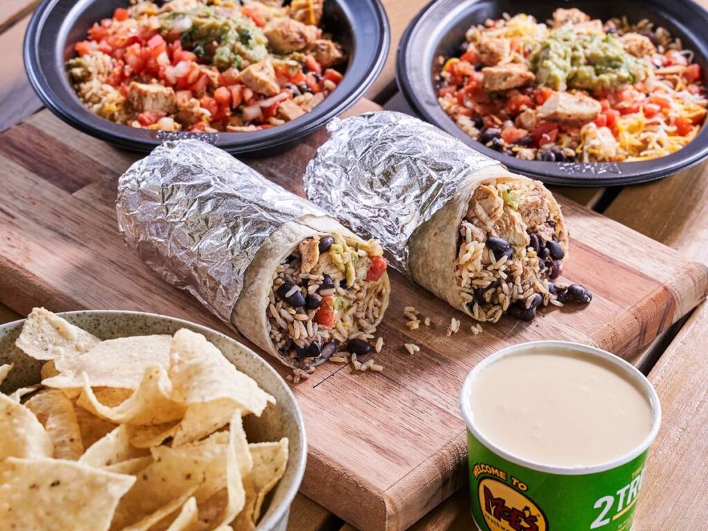 Two foil-wrapped burritos on a board, with two black bowls of mixed rice and chicken, a bowl of tortilla chips, and a cup of queso dip.