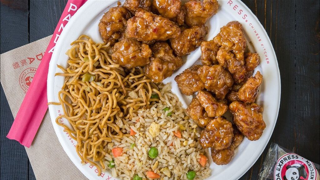 A plate of orange chicken, chow mein, and fried rice from Panda Express on a table with napkins and chopsticks.