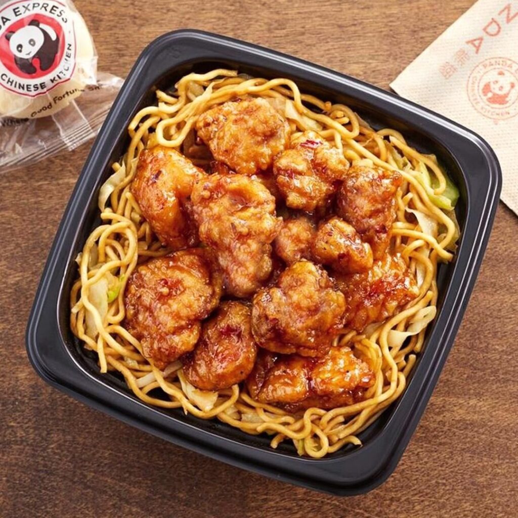 A black container filled with noodles topped with glazed fried chicken pieces. A soy sauce packet and napkin are placed beside the container on a wooden surface.