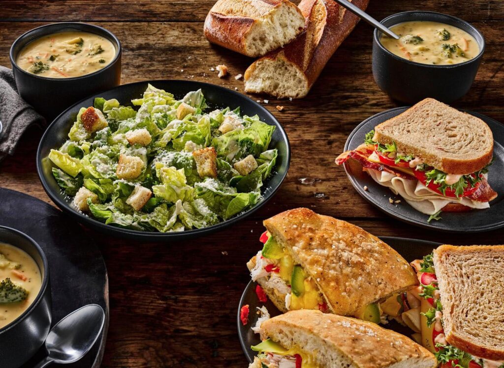 Assorted sandwiches, Caesar salad, and bowls of soup on a wooden table with crusty bread.