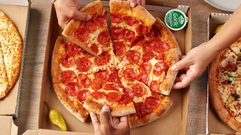 Three people grabbing slices from a pepperoni pizza in a box, with a small garlic sauce cup nearby.