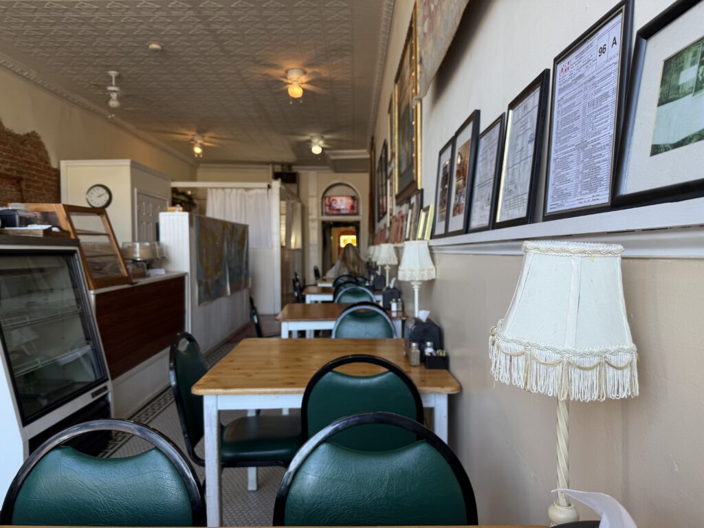 Interior of a quaint cafe with green cushioned chairs, wooden tables, a display case, framed pictures on the wall, and a lace-shaded lamp.