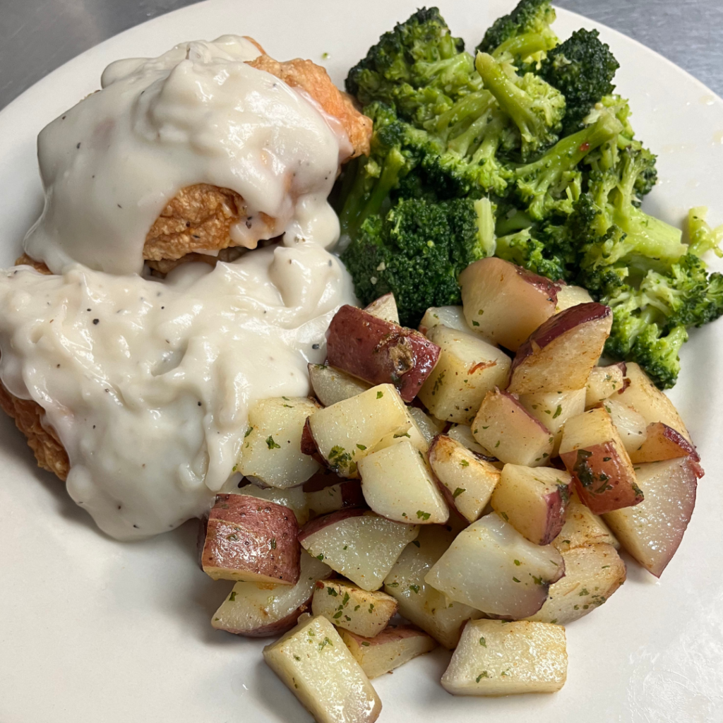 A plate with roasted potatoes, broccoli, and fried chicken topped with creamy white gravy.