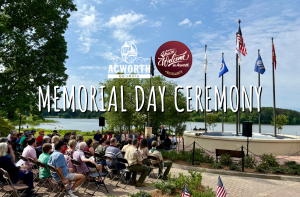 People sit outdoors by a lake, facing a stage with five raised flags, attending a Memorial Day ceremony in Acworth, Georgia.