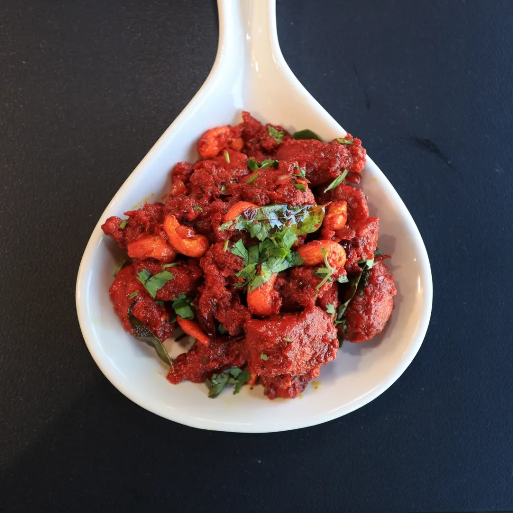 A white spoon-shaped dish holds a serving of bright red curry with visible cashews and herbs, set on a black surface.