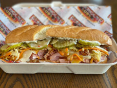 A sub sandwich with ham, cheese, pickles, shredded vegetables, and dressing in a takeout container on a table.