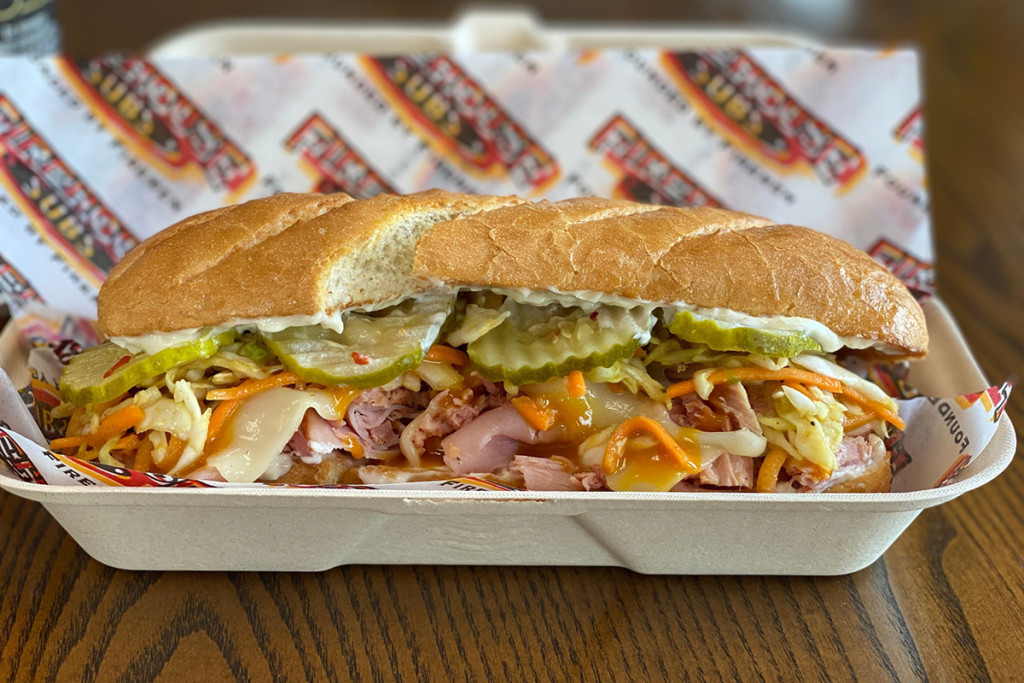 A sub sandwich with ham, cheese, pickles, shredded vegetables, and dressing in a takeout container on a table.