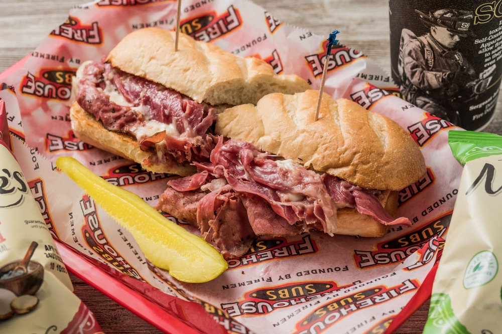 Sub sandwich with sliced meat and melted cheese on a tray, alongside a pickle spear and chip bags.