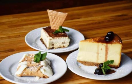 Three plates of desserts on a wooden surface: a slice of cheesecake with cherries, a piece of tiramisu garnished with a wafer, and two cannoli filled with creamy filling, dusted with powdered sugar.