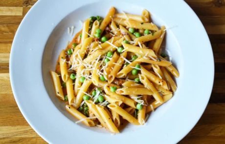 A white plate filled with penne pasta mixed with green peas and topped with grated cheese, placed on a wooden table.