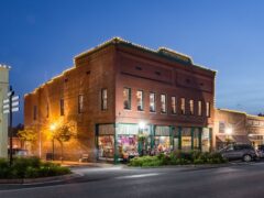 Historic brick building with lit outline at dusk, street corner, windows lit, parked cars, and street signs visible.