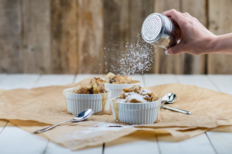 A hand sprinkles powdered sugar over three ramekins of dessert on a wooden table, with two spoons beside them.