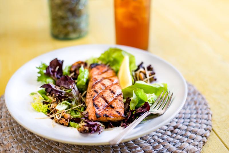 A grilled salmon fillet atop a mixed green salad with a lemon wedge on a white plate, placed on a woven mat. A fork lies on the plate edge. An iced tea is in the background.