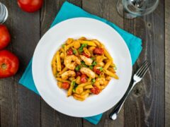 A plate of penne pasta with shrimp, cherry tomatoes, and garnished with green onions is on a teal napkin. Fork and glass beside it, with two red tomatoes in the background.