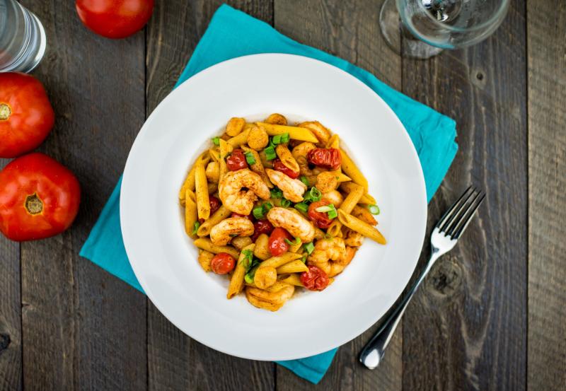 A plate of penne pasta with shrimp, cherry tomatoes, and garnished with green onions is on a teal napkin. Fork and glass beside it, with two red tomatoes in the background.