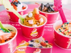 Five cups of colorful frozen yogurt with various toppings, including fruit, sprinkles, and candy, on a pink surface.