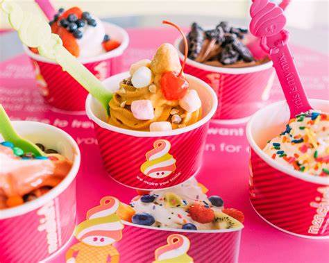 Five cups of colorful frozen yogurt with various toppings, including fruit, sprinkles, and candy, on a pink surface.