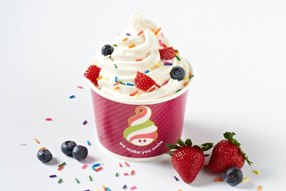 A cup of frozen yogurt with blueberries, strawberry slices, and rainbow sprinkles. Two whole strawberries and blueberries are placed beside the cup.