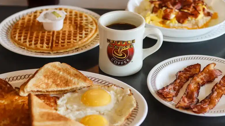 A table with a breakfast spread: eggs, toast, bacon, waffles with butter, and a mug of coffee.