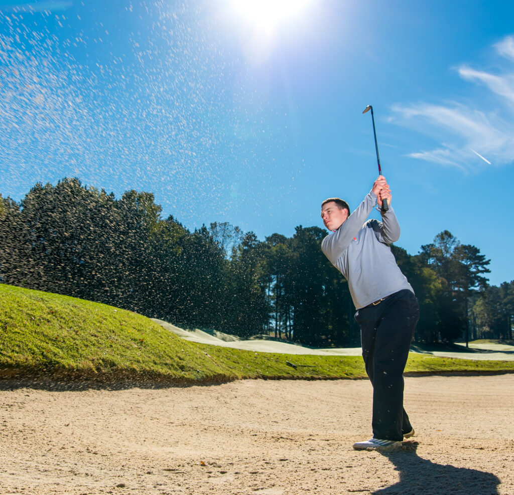 A golfer wearing a gray sweater hits a shot from a sand bunker on a sunny day with greenery and trees in the background.