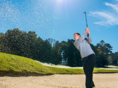 A golfer wearing a gray sweater hits a shot from a sand bunker on a sunny day with greenery and trees in the background.