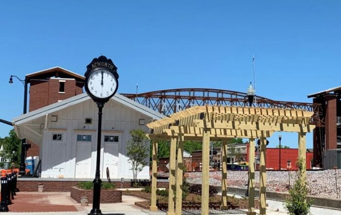 Outdoor clock and wooden pergola structure near a railroad track with a brick building and a pedestrian bridge in the background on a sunny day.