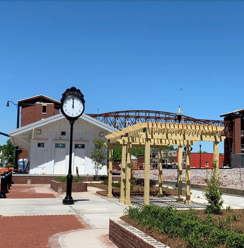 Outdoor clock and wooden pergola structure near a railroad track with a brick building and a pedestrian bridge in the background on a sunny day.