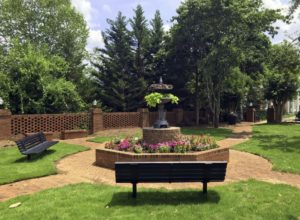 A small park with brick pathways, a central decorative fountain surrounded by flowerbeds, and black benches. Trees and a brick wall are in the background under a partly cloudy sky.