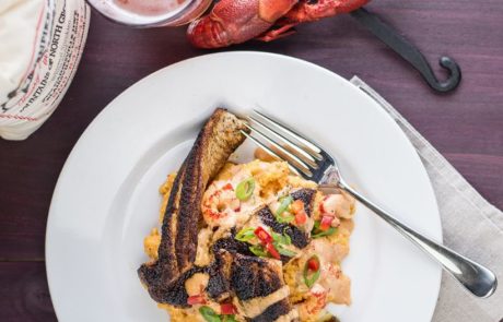 A plate of blackened fish atop a creamy crawfish dish with a fork on the side. A drink, a lobster, and a lemon are positioned nearby on a wooden surface.