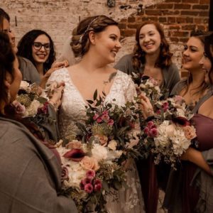 A bride, wearing a white lace gown and holding a bouquet, smiles while standing with bridesmaids dressed in gray shawls and purple dresses, all holding floral bouquets, against a brick wall background.