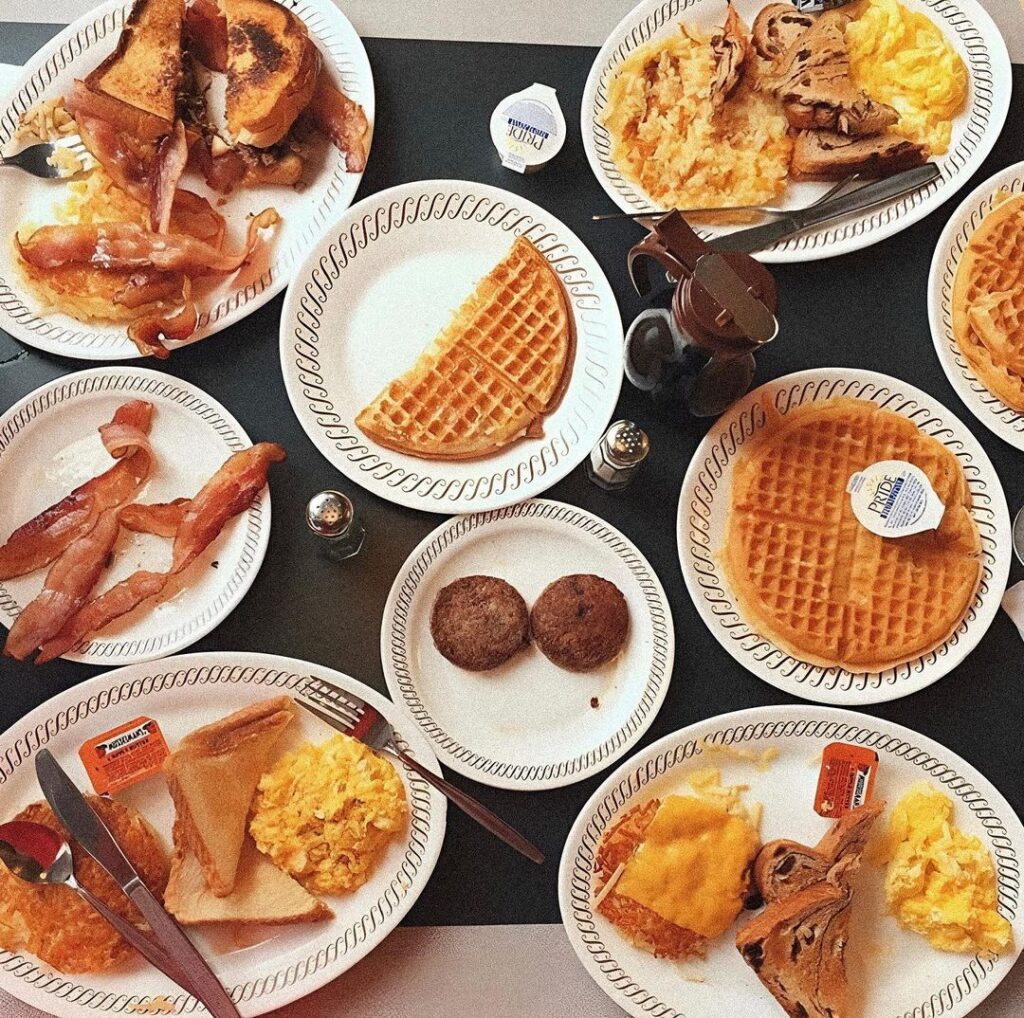 A table with plates of waffles, scrambled eggs, bacon, sausage, toast, and syrup packets.